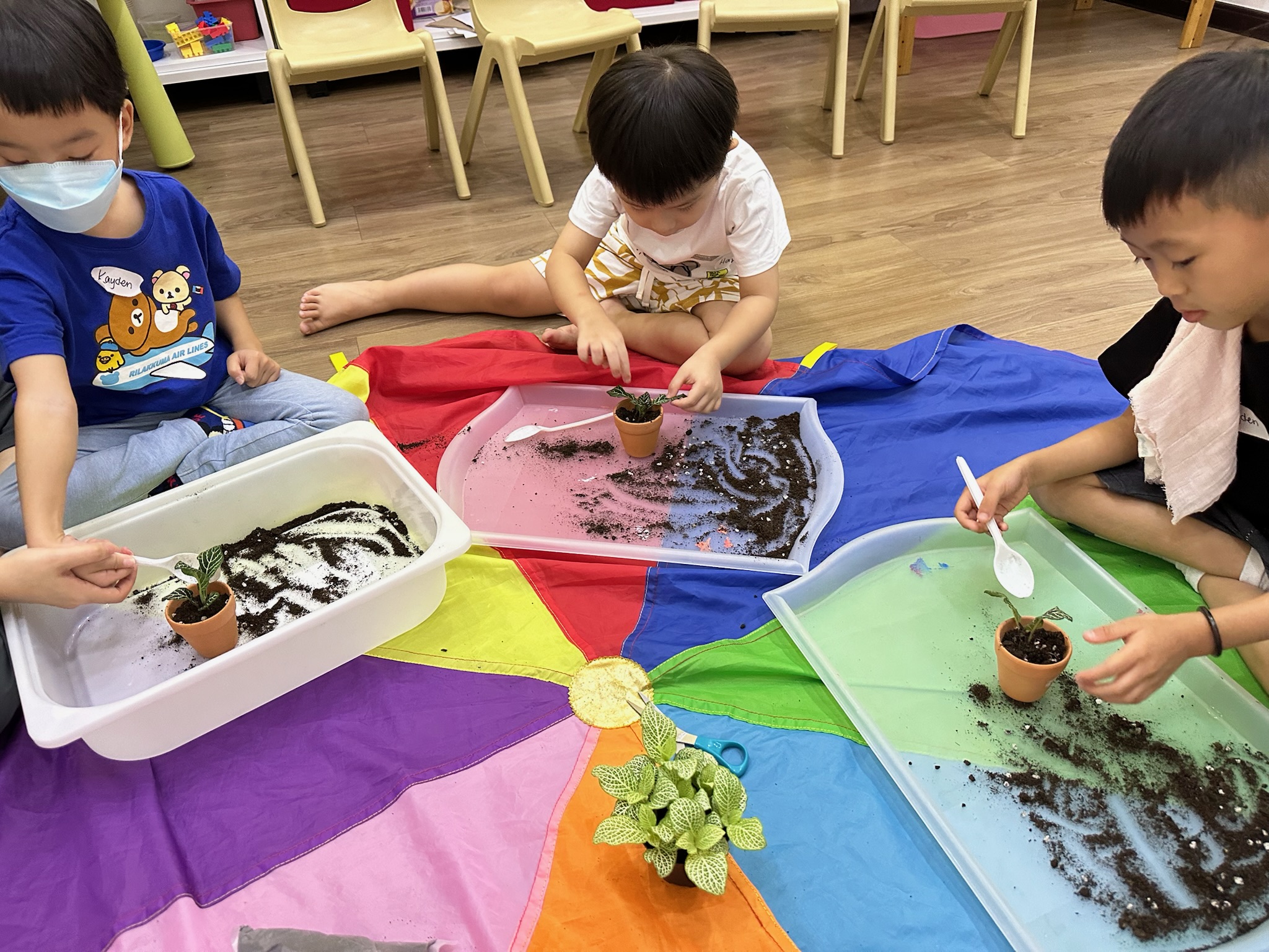 Three children potting small plants at separate trays on a rainbow parachute, using spoons to move soil