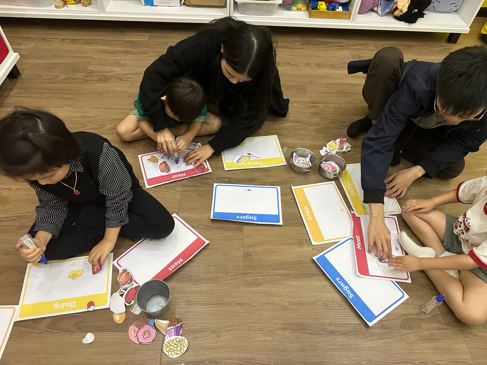 Children and adults sitting on the floor gluing food pictures onto labelled cards for meat, dairy, grain and sugars