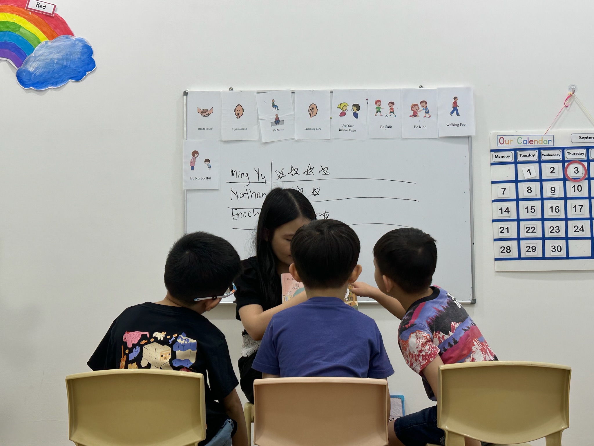 Three children seated in small chairs with a teacher in front of a whiteboard showing picture cards for classroom rules and a star chart