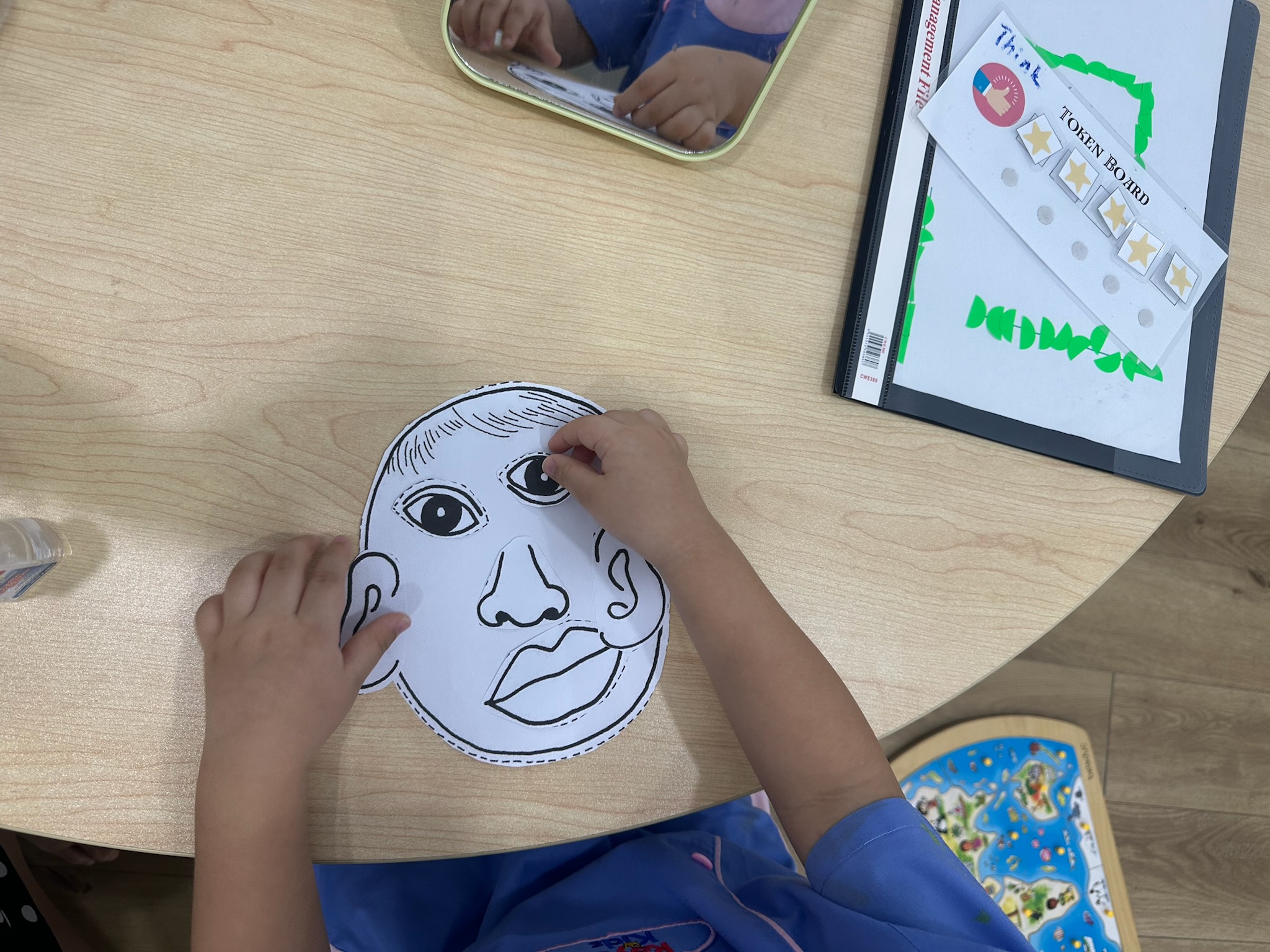 A child&rsquo;s hands placing cut-out eyes, nose and mouth onto a paper face, with a mirror and a token board on the table