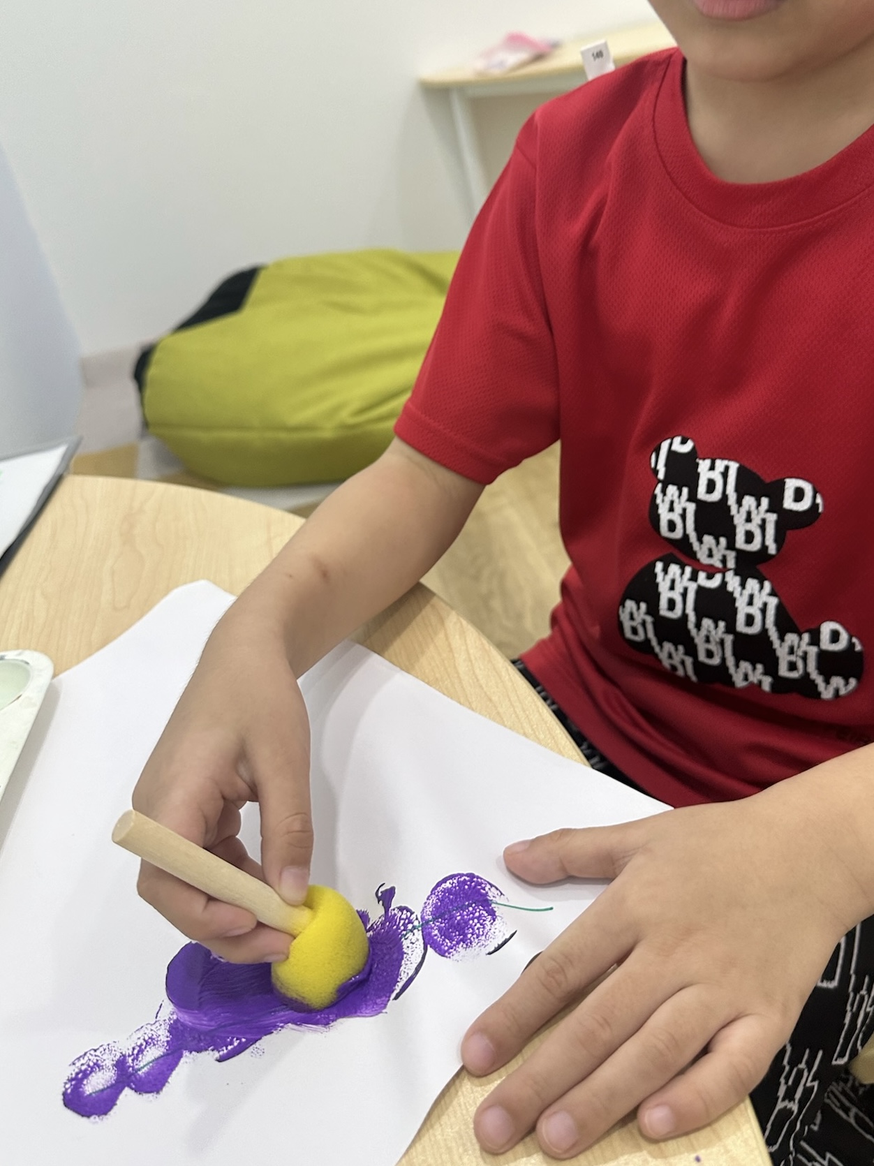 A child concentrating while painting purple shapes on paper with a yellow sponge brush