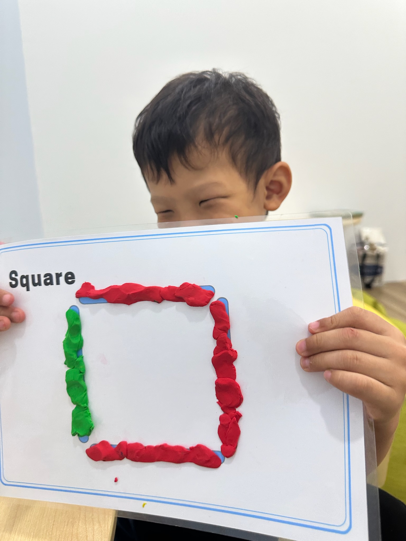 A smiling child holding up a laminated card with a square made from red and green play dough